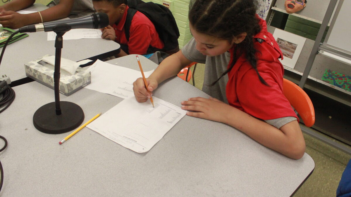Two youth writing on pieces of paper sitting behind a microphone in a classroom