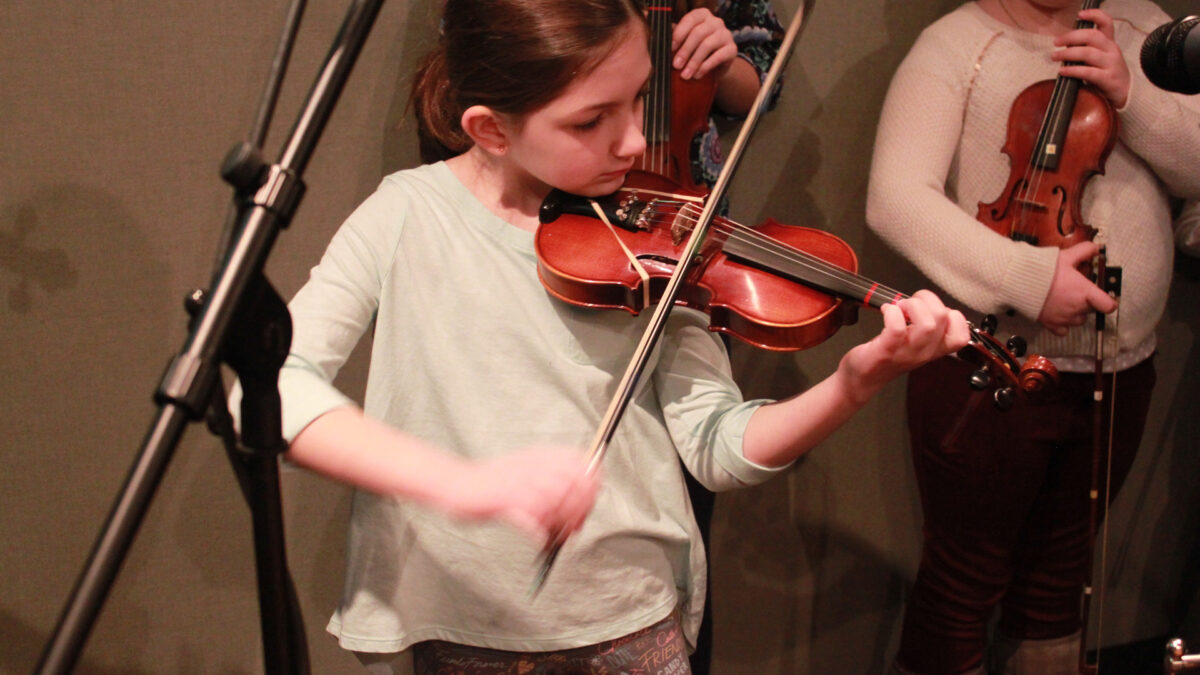 A youth playing a violin and two youth holding violins standing in the background in a radio studio