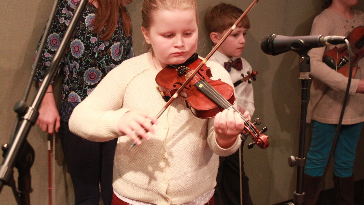 A youth playing a violin and three youth holding violins standing in the background in a radio studio