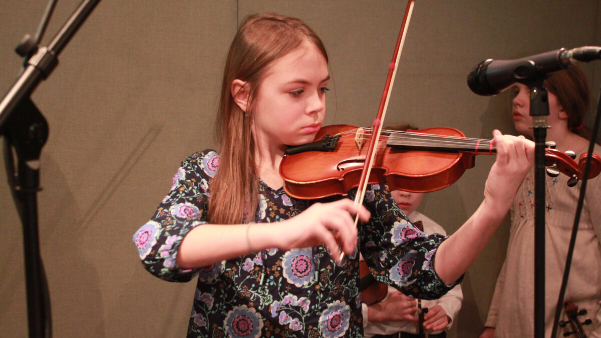 A youth playing a violin and two youth holding violins standing in the background in a radio studio