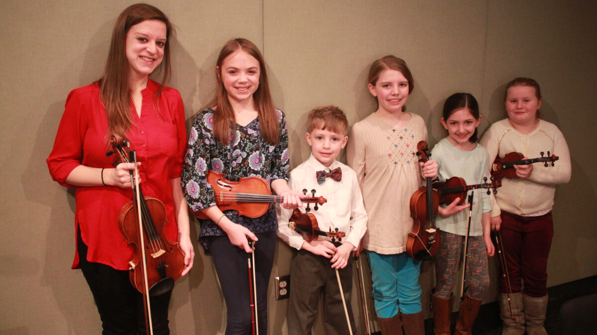 An adult and five youth smiling and holding violins in a radio studio