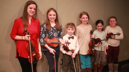 An adult and five youth smiling and holding violins in a radio studio