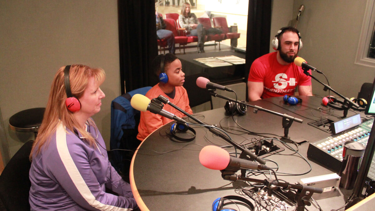 Two adults and a youth wearing headphones speaking into microphones in a radio studio