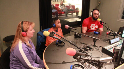 Two adults and a youth wearing headphones speaking into microphones in a radio studio