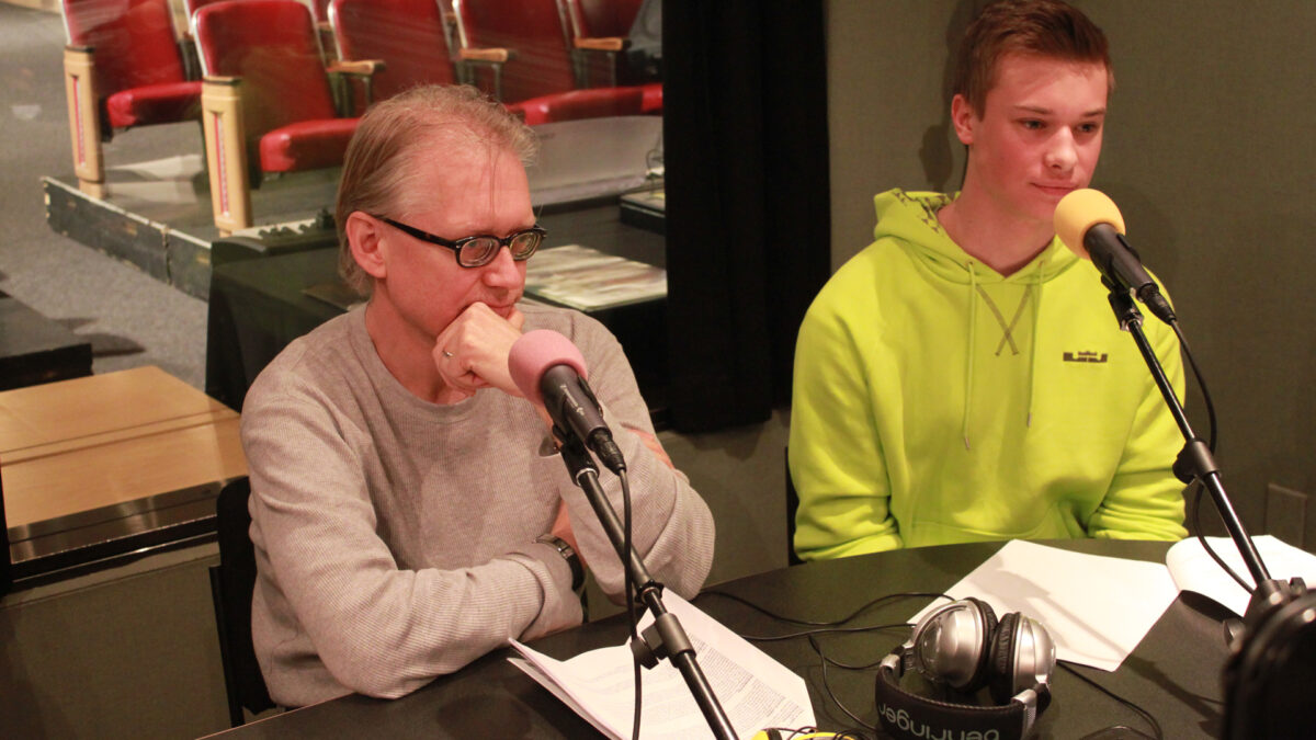 A teen and an adult sitting behind microphones and pieces of paper in a radio studio