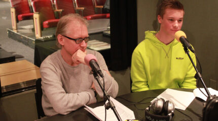 A teen and an adult sitting behind microphones and pieces of paper in a radio studio