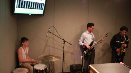 A teen playing a drum set, a teen playing electric bass, and a teen playing alto saxophone in a radio studio