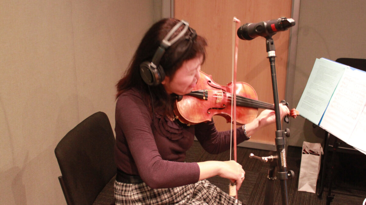An adult wearing headphones playing violin and looking at sheet music on a music stand in a radio studio