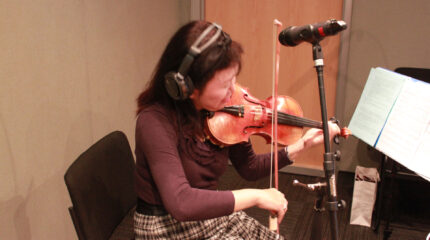 An adult wearing headphones playing violin and looking at sheet music on a music stand in a radio studio