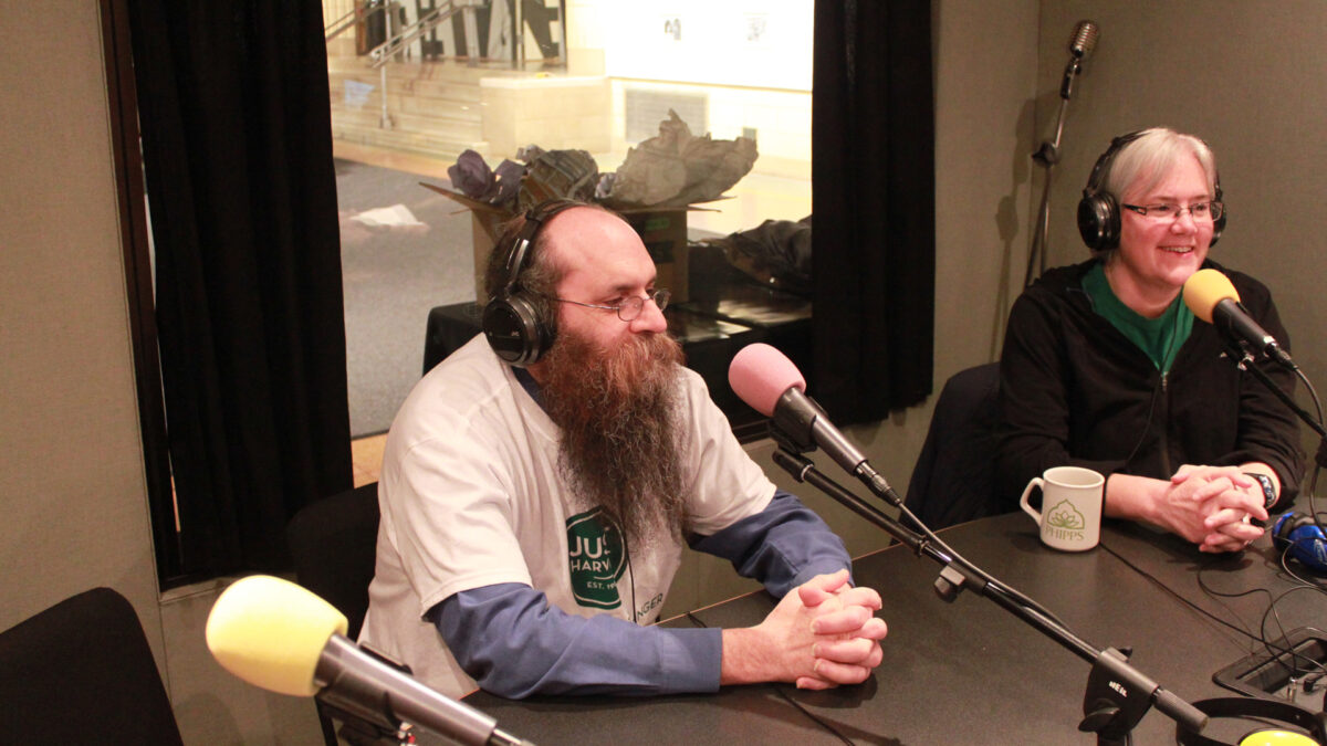 Two adults wearing headphones speaking into microphones in a radio studio