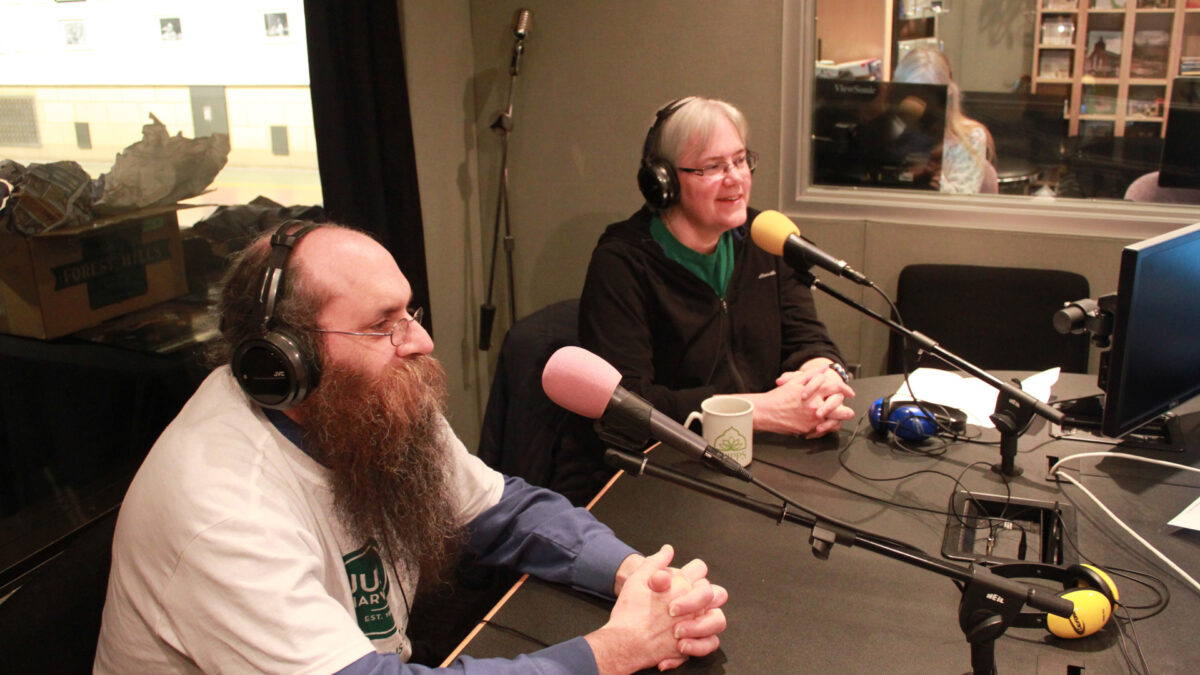 Two adults wearing headphones speaking into microphones in a radio studio