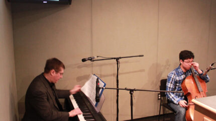 A youth playing a cello and an adult playing a piano keyboard sitting behind a microphone in a radio studio