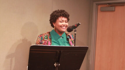 A teen smiling standing behind a microphone and a music stand in a radio studio