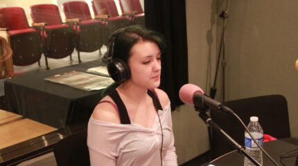 A teen wearing headphones speaking into a microphone in a radio studio