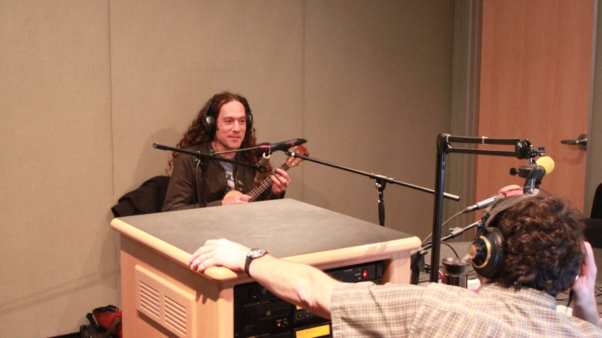 An adult wearing headphones playing a ukulele as another adult wearing headphones listens in a radio studio