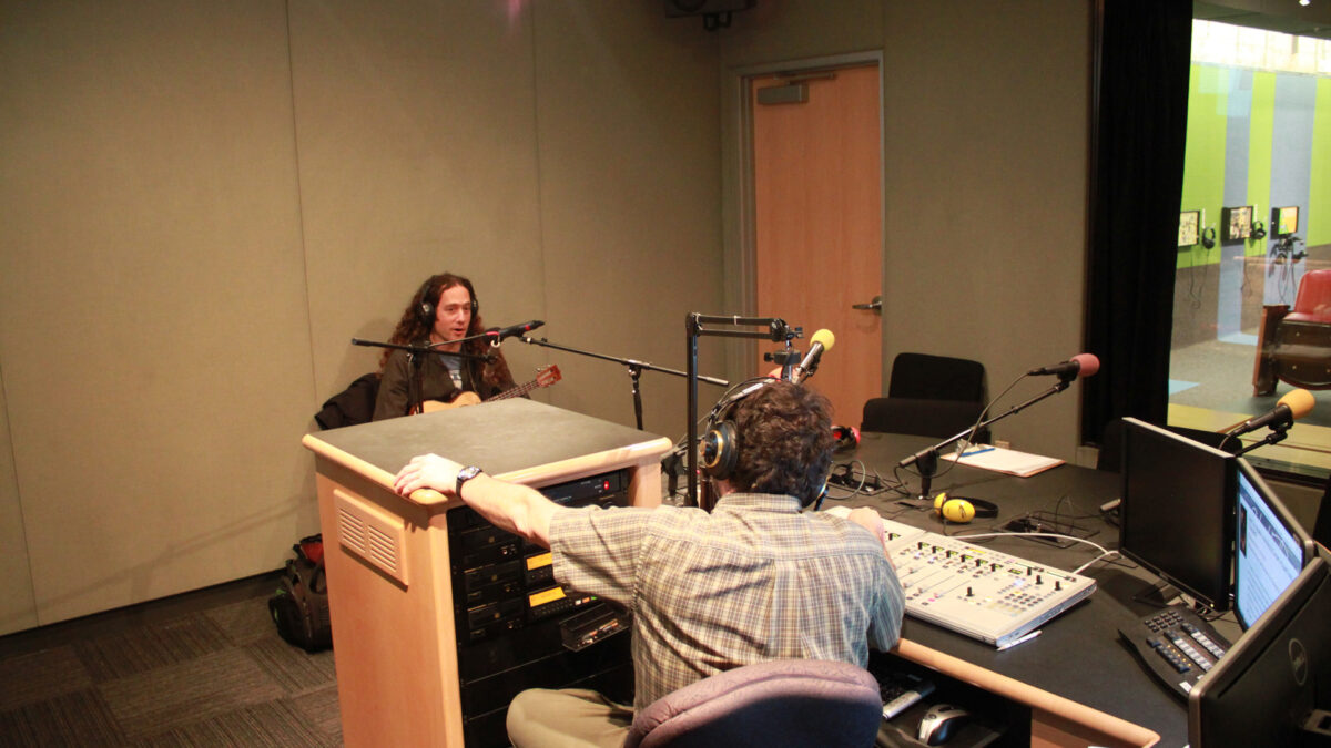 An adult wearing headphones playing ukulele and singing into a microphone as an adult wearing headphones sitting behind a microphone and a mixer listens in a radio studio
