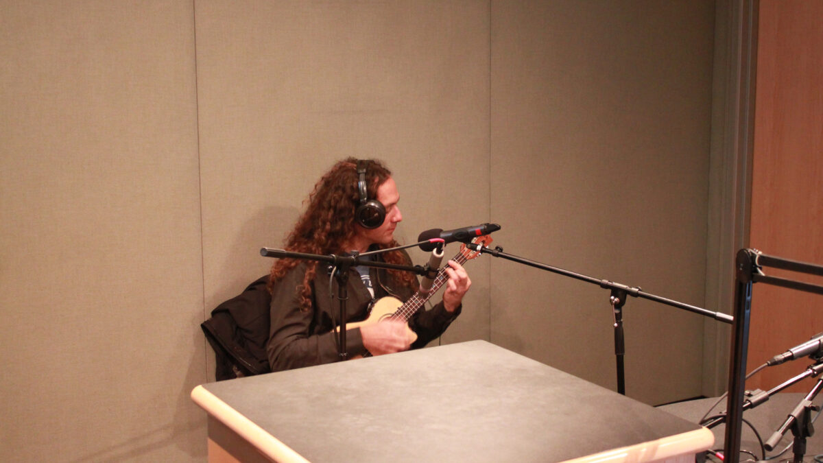 An adult wearing headphones playing ukulele and sitting behind a microphone in a radio studio