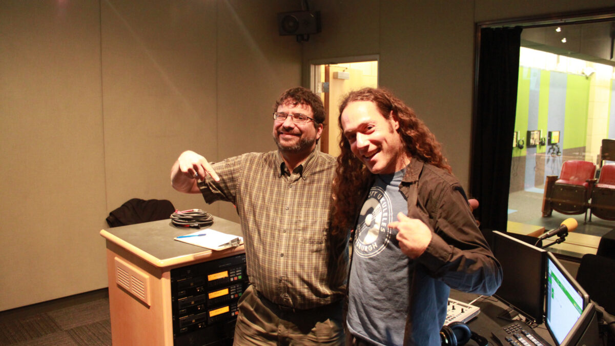 Two adults smiling and posing with a finger pointing downwards in a radio studio