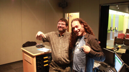 Two adults smiling and posing with a finger pointing downwards in a radio studio