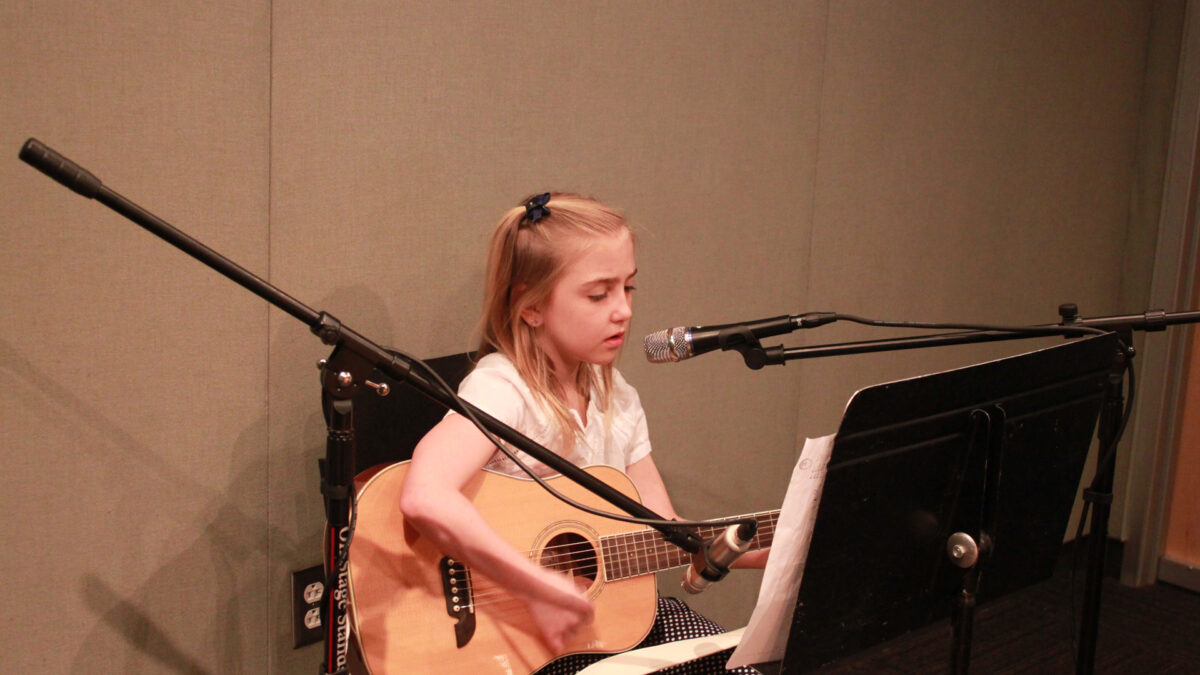 A youth playing an acoustic guitar and singing into a microphone looking at sheet music on a music stand in a radio studio