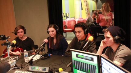 Four teens wearing headphones and speaking into microphones being interviewed in a radio studio