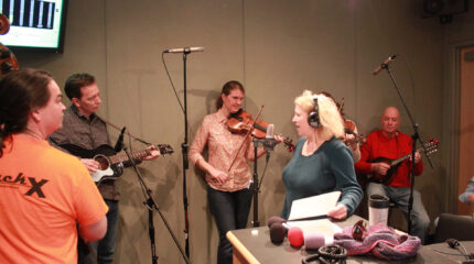 An adult playing an upright bass, an adult playing an acoustic guitar, two adults playing violins, an adult playing a mandolin, and an adult wearing headphones singing into a microphone in a radio studio