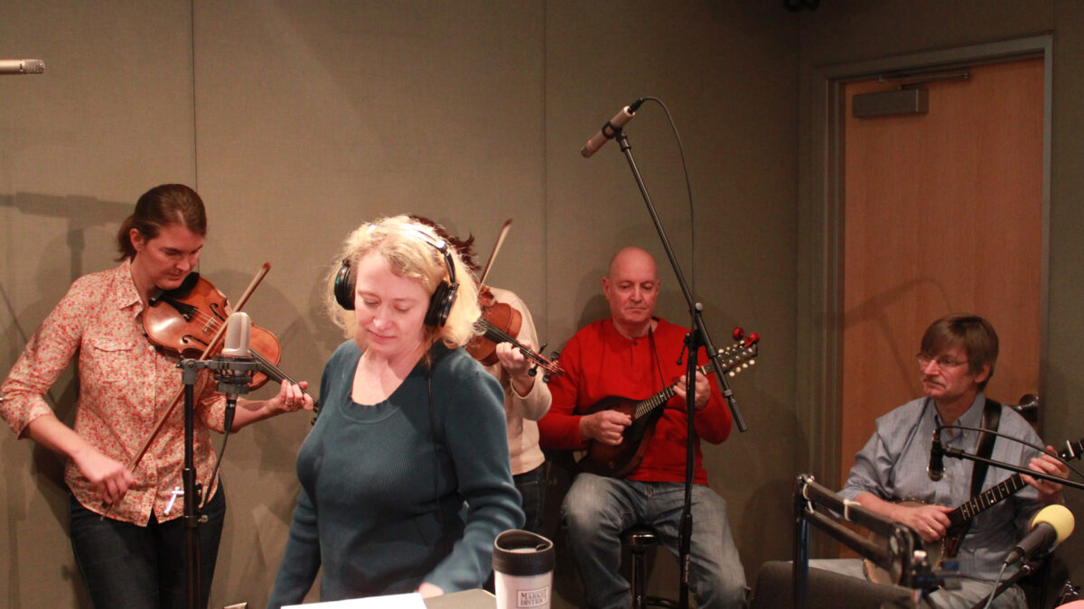 Two adults playing violins, an adult playing mandolin, an adult playing banjo, and an adult wearing headphones looking down at a piece of paper in a radio studio