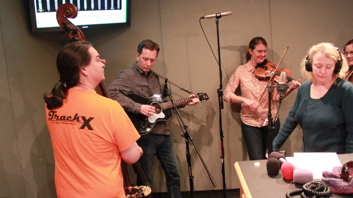 An adult playing upright bass, an adult playing acoustic guitar, an adult playing a violin, and an adult wearing headphones looking down at a piece of paper in a radio studio