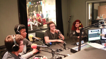 Three teens and an adult wearing headphones and speaking into microphones in a radio studio