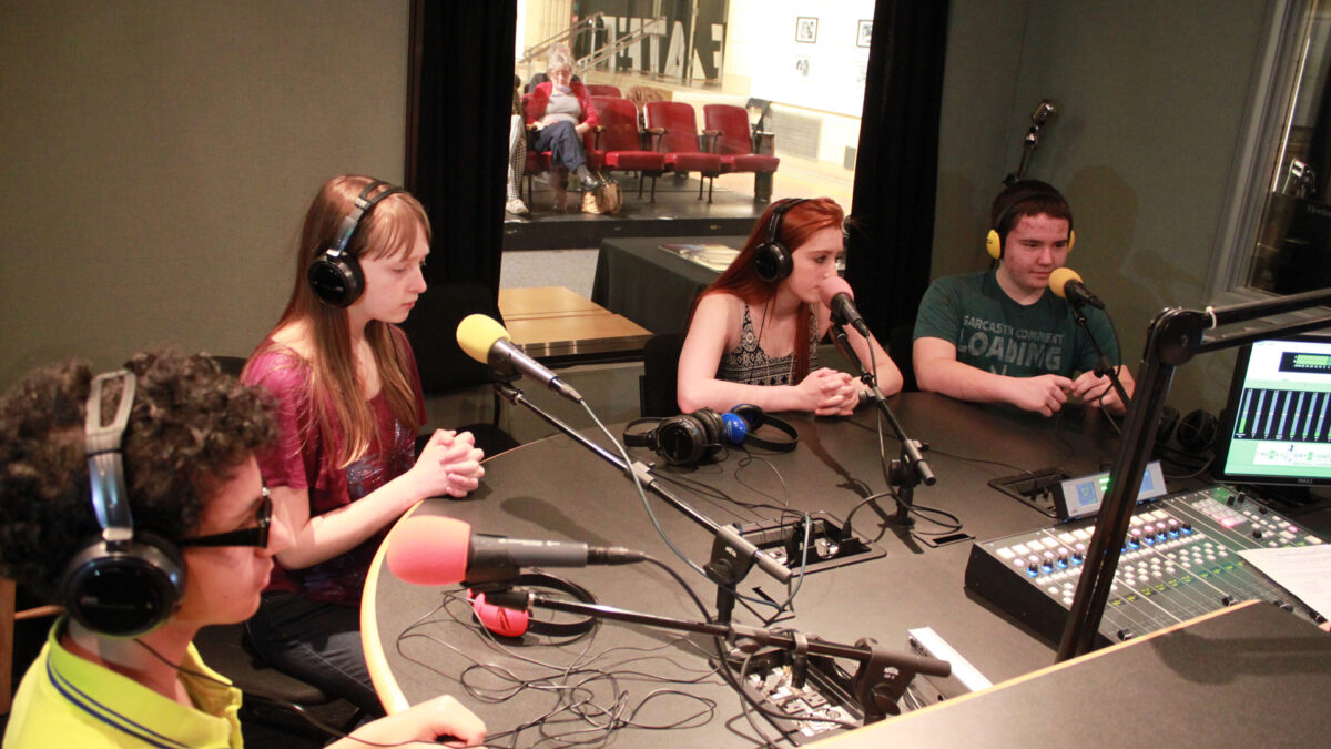 Four teens wearing headphones speaking into microphones in a radio studio