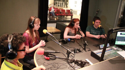 Four teens wearing headphones speaking into microphones in a radio studio