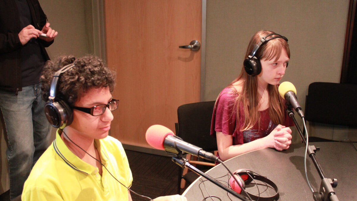 Two teens wearing headphones speaking into microphones in a radio studio
