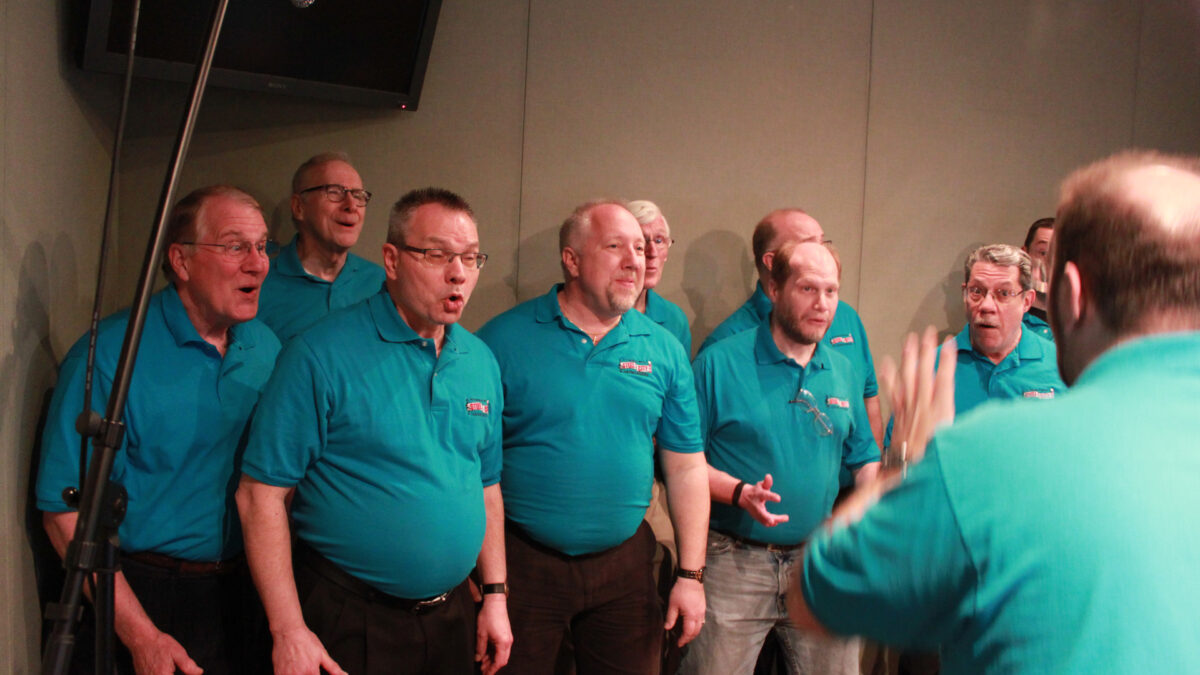 An adult conducting nine other adults singing in a choir in a radio studio