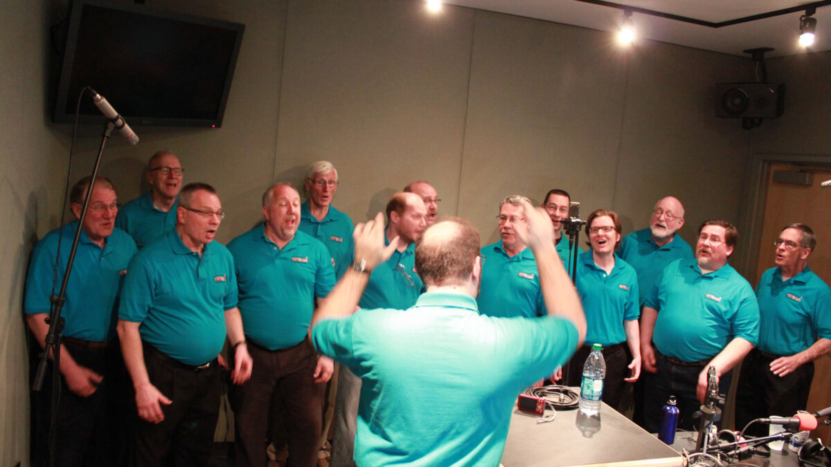 An adult conducting thirteen other adults singing in a choir in a radio studio
