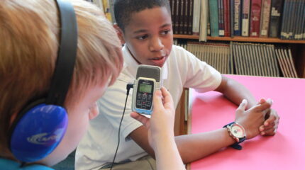 A youth wearing headphones interviewing another youth with a portable recorder
