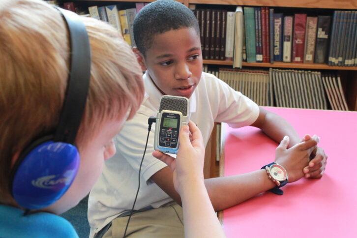 A youth wearing headphones interviewing another youth with a portable recorder