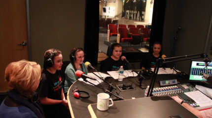 Five youth wearing headphones speaking into microphones in a radio studio
