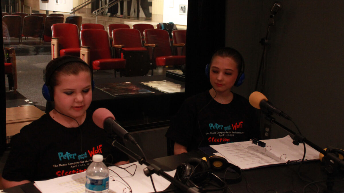 IMG_8083_wp Two youth wearing headphones speaking into microphones looking down at scripts in a radio studio