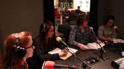 Four teens wearing headphones speaking into microphones in a radio studio