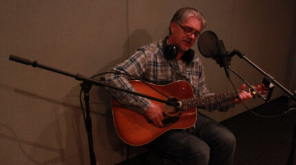 An adult playing an acoustic guitar and singing into a microphone in a radio studio