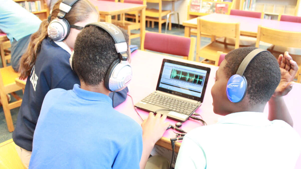 Three youth wearing headphones looking at audio editing software on a laptop computer in a library