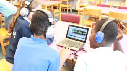 Three youth wearing headphones looking at audio editing software on a laptop computer in a library