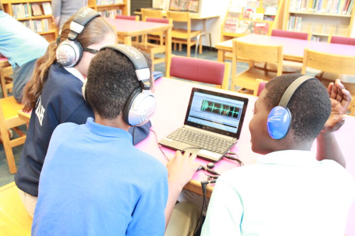Three youth wearing headphones looking at audio editing software on a laptop computer in a library