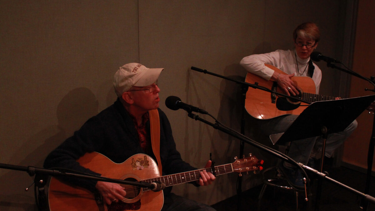 Two adults playing acoustic guitars and singing into microphones in a radio studio