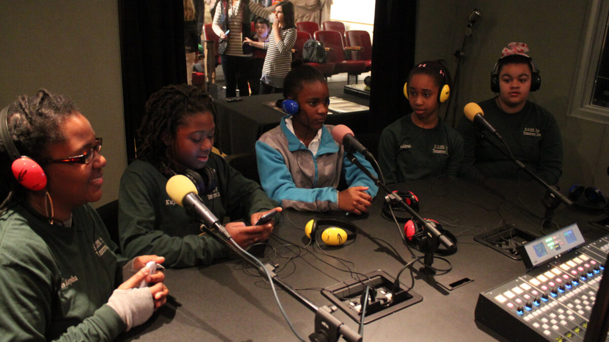 Five teens wearing headphones speaking into microphones in a radio studio