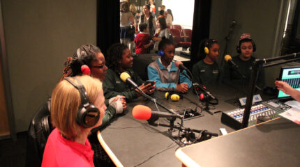 Four youth and two adults wearing headphones speaking into microphones in a radio studio