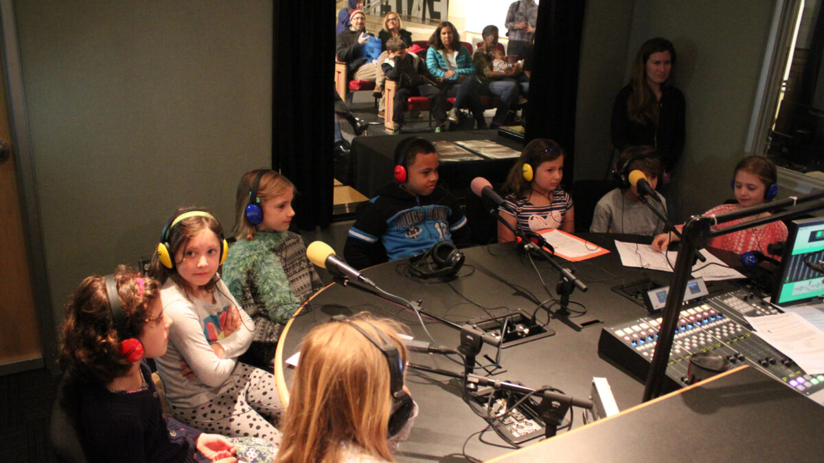 IMG_8462_wp Eight youth wearing headphones speaking into microphones as an adult watches from a corner in a radio studio