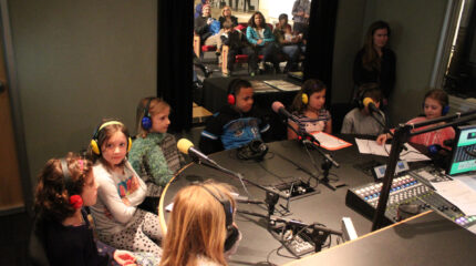 Eight youth wearing headphones speaking into microphones as an adult watches from a corner in a radio studio