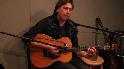 An adult playing an acoustic guitar and singing into a microphone in a radio studio