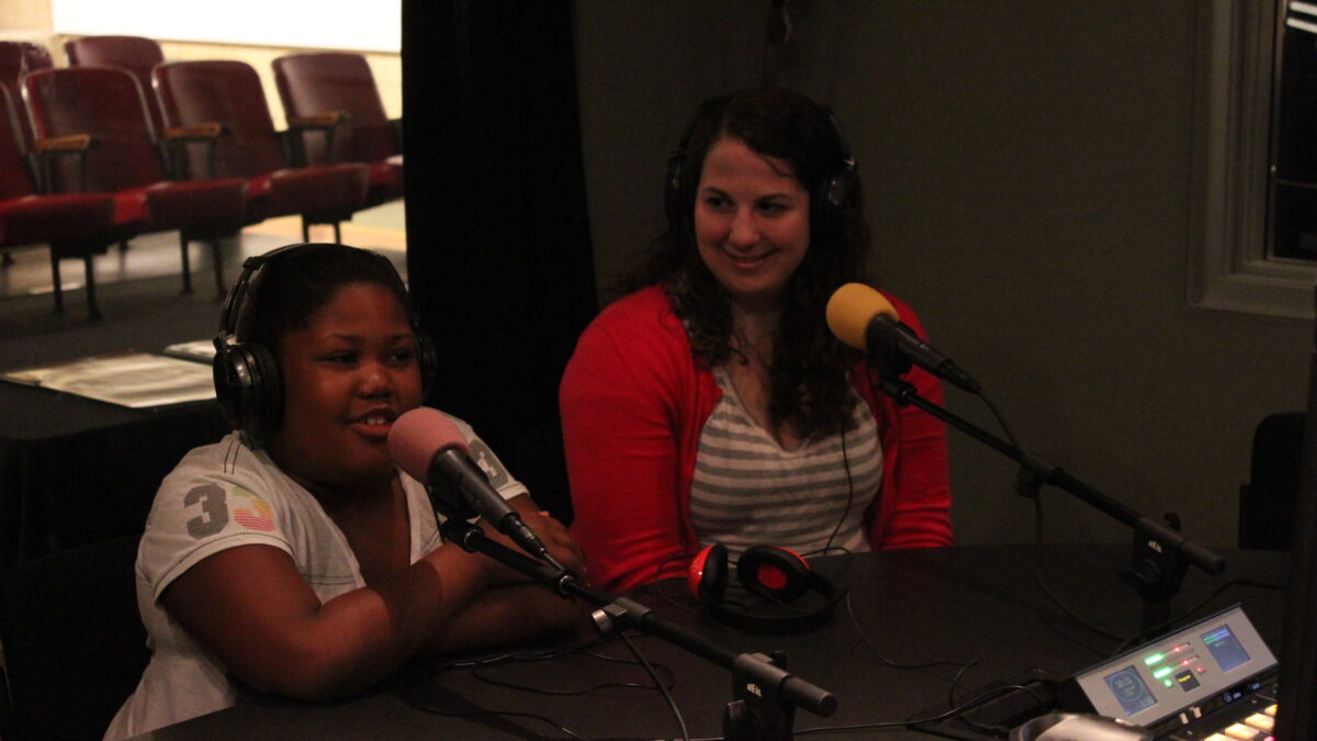 An adult and a youth wearing headphones speaking into microphones in a radio studio
